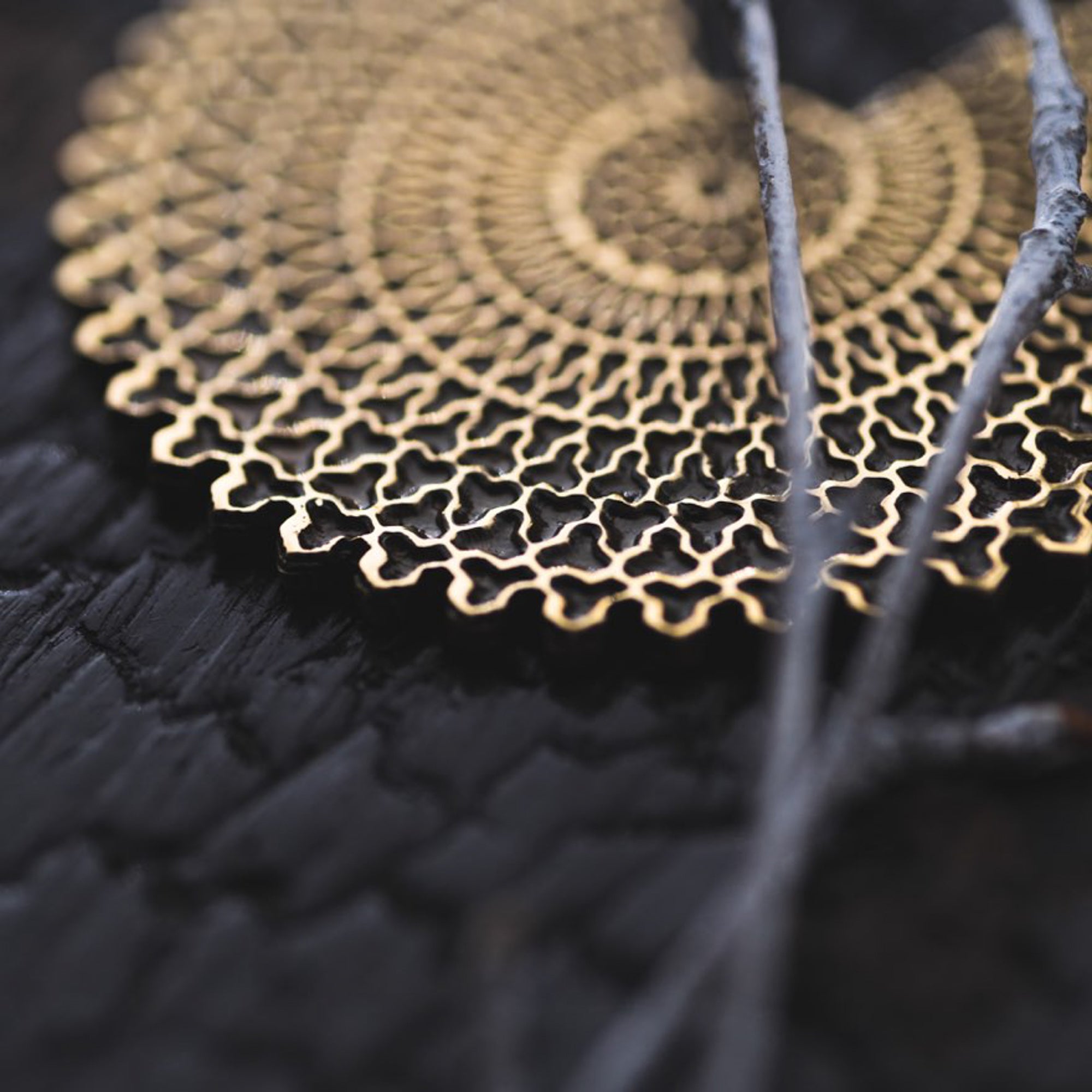 A close-up of an intricately patterned, circular golden object on a dark, textured surface. The object resembles the VORTEX Tribal Oversized Mandala Disc Hoop Earrings in Gold by Kaya Movement, featuring a repeating geometric design and is partially covered by several thin, gray branches in the foreground.